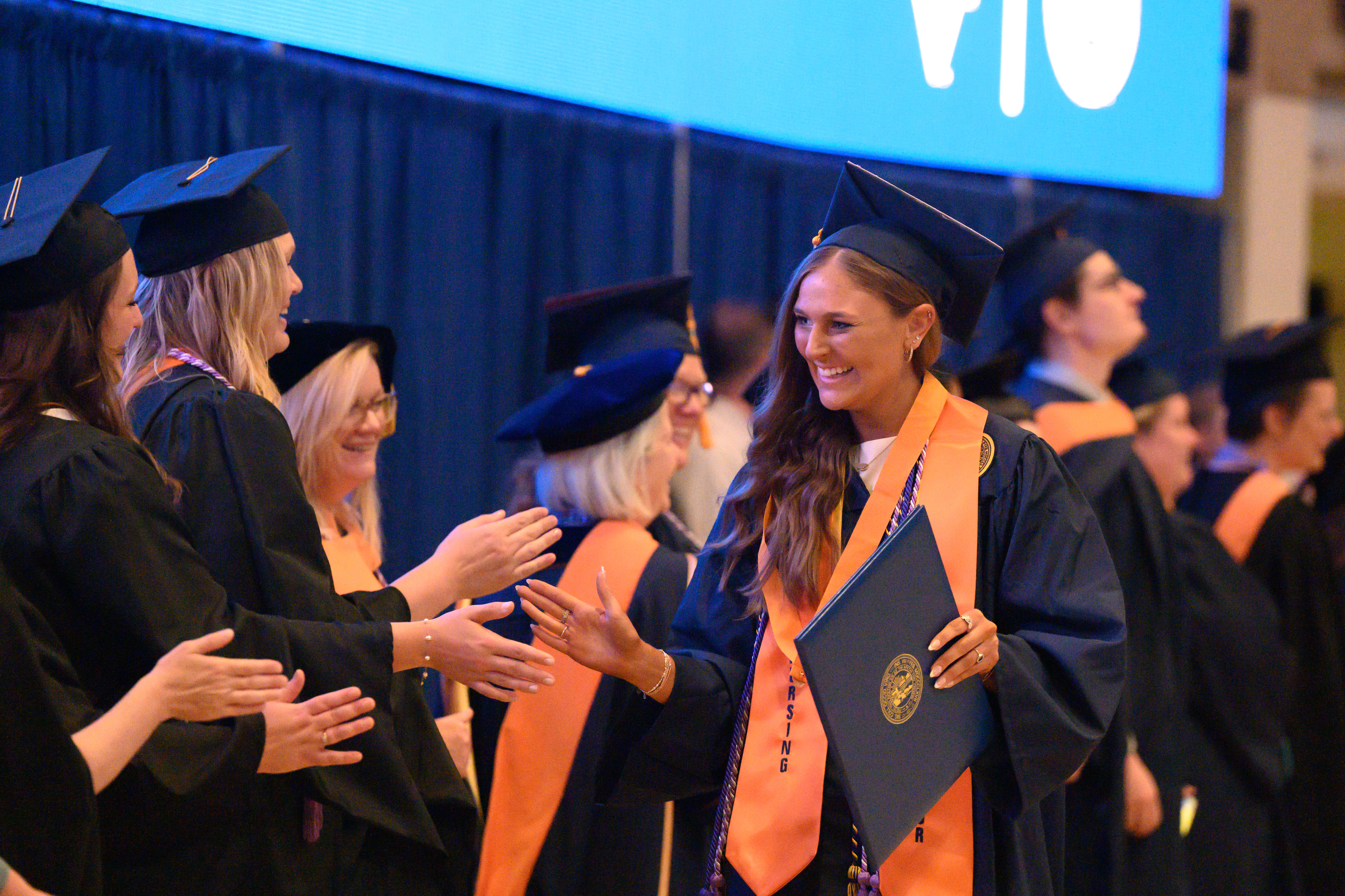 A graduating nursing student walks past other graduating students, high fiving them. 